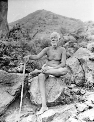 Photo of Ramana Maharshi sat on rock with cane arunachala behind dakshinamurthy pose in black and white