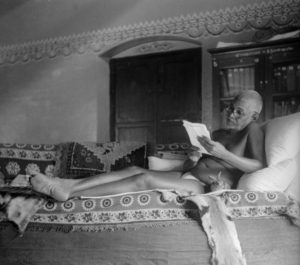 Photo of Ramana Maharshi seated couch in old hall correcting rabbit at side in black and white